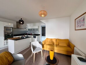 A modern kitchen with white cabinets, a black countertop, and stainless steel appliances. There is a yellow sofa and a gray chair in the living area. - Modern duplex for 5, ocean view, with wifi (Saint-Hilaire-de-Riez)