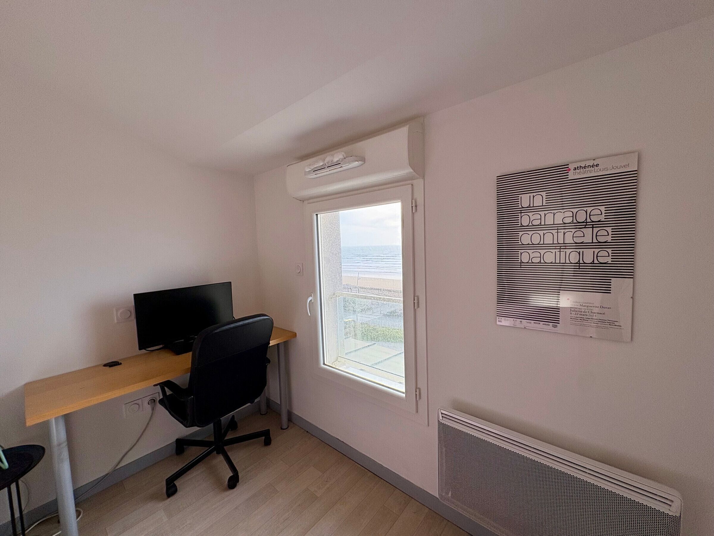 A small office space with a desk, chair, and a computer. There is a window with a view of the sea and a poster on the wall.