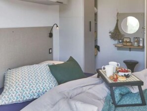 Una cama bien hecha con una almohada estampada, una mesita de noche con una bandeja de desayuno y un espejo sobre una repisa con una planta.
