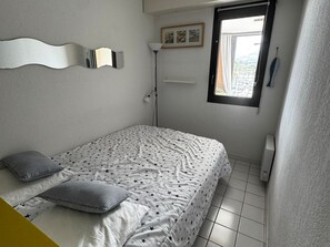 A bedroom with a bed, pillows, a wall-mounted mirror, a window with a view, and a radiator. - Air-Conditioned Apartment with Private Parking - Argelès-sur-Mer - 4 people (Argelès-sur-Mer)