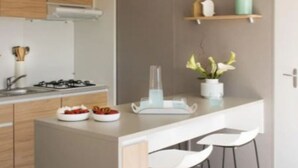 A modern kitchen with wooden cabinets, a gas stove, a white countertop, and a bar area with white bar stools. - Mobile home for 4 in Saint-Jean-de-Monts (Saint-Jean-de-Monts)
