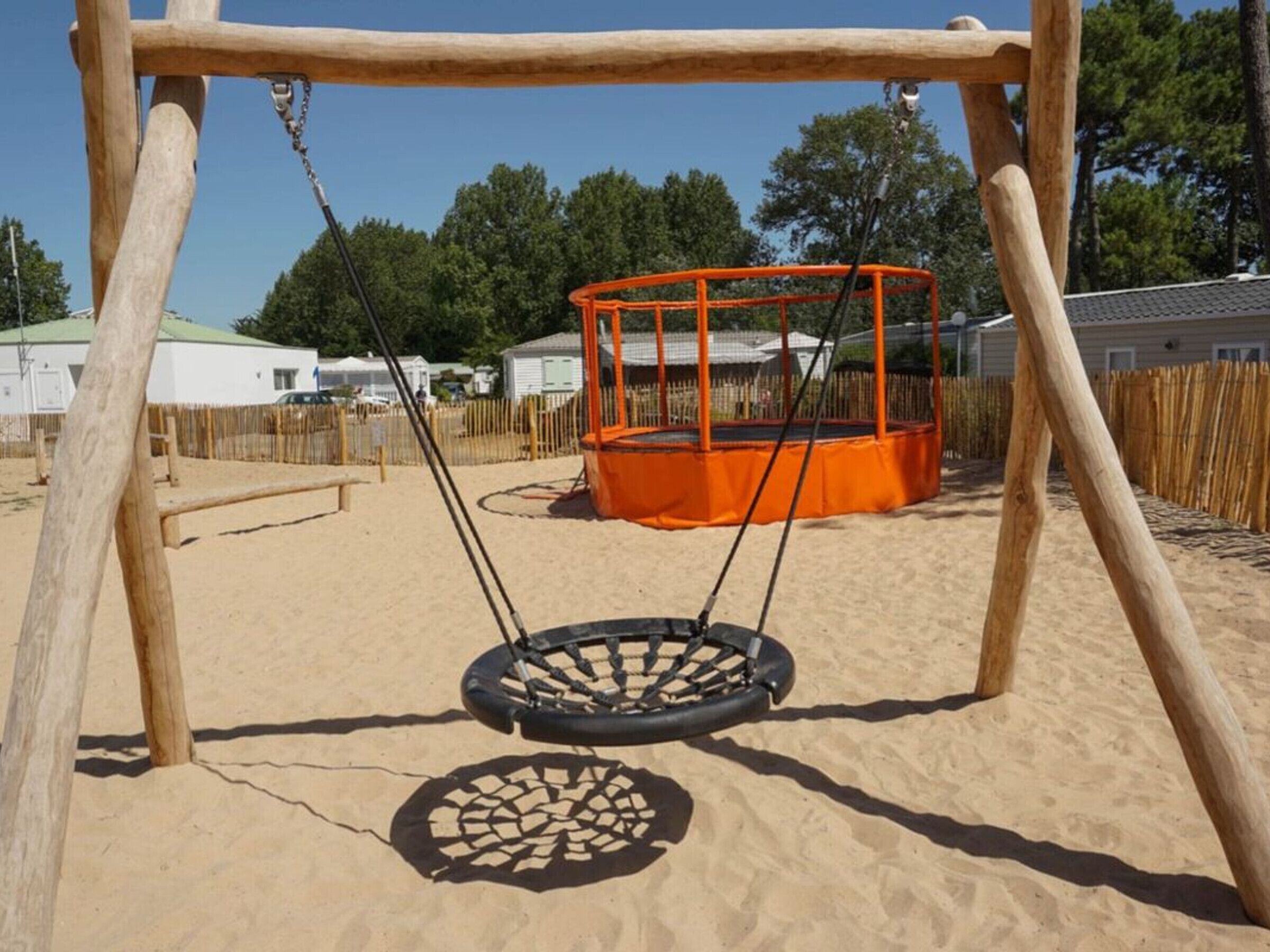 A wooden swing set with a tire swing, a fenced play area, and a building in the background.