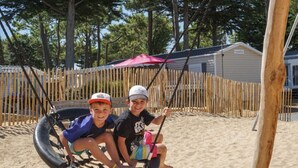 Two people on a swing set in a sandy area with a wooden fence and a building in the background. - Mobile home for 4 in Saint-Jean-de-Monts (Saint-Jean-de-Monts)