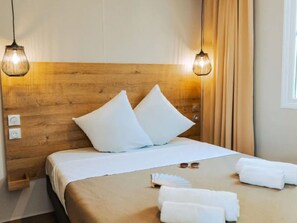 Et hotelværelse med sengegavl i træ, to hvide puder, en pænt redt seng med hvide sengetøj, et par solbriller og et foldet hvidt håndklæde på sengen.
