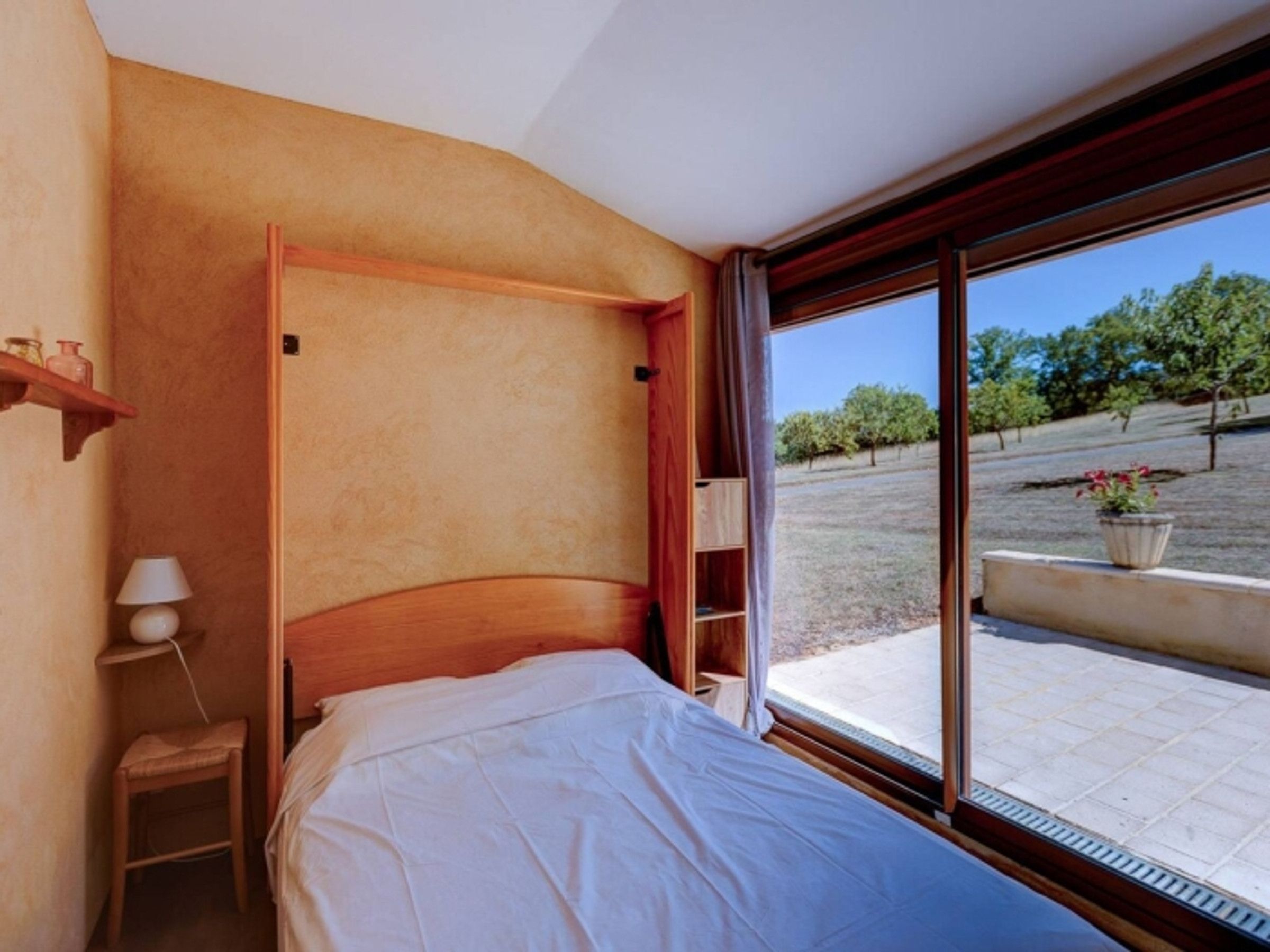 Un dormitorio con cama de madera, una mesita de noche, una lámpara y un amplio ventanal con vistas a árboles y un jardín.