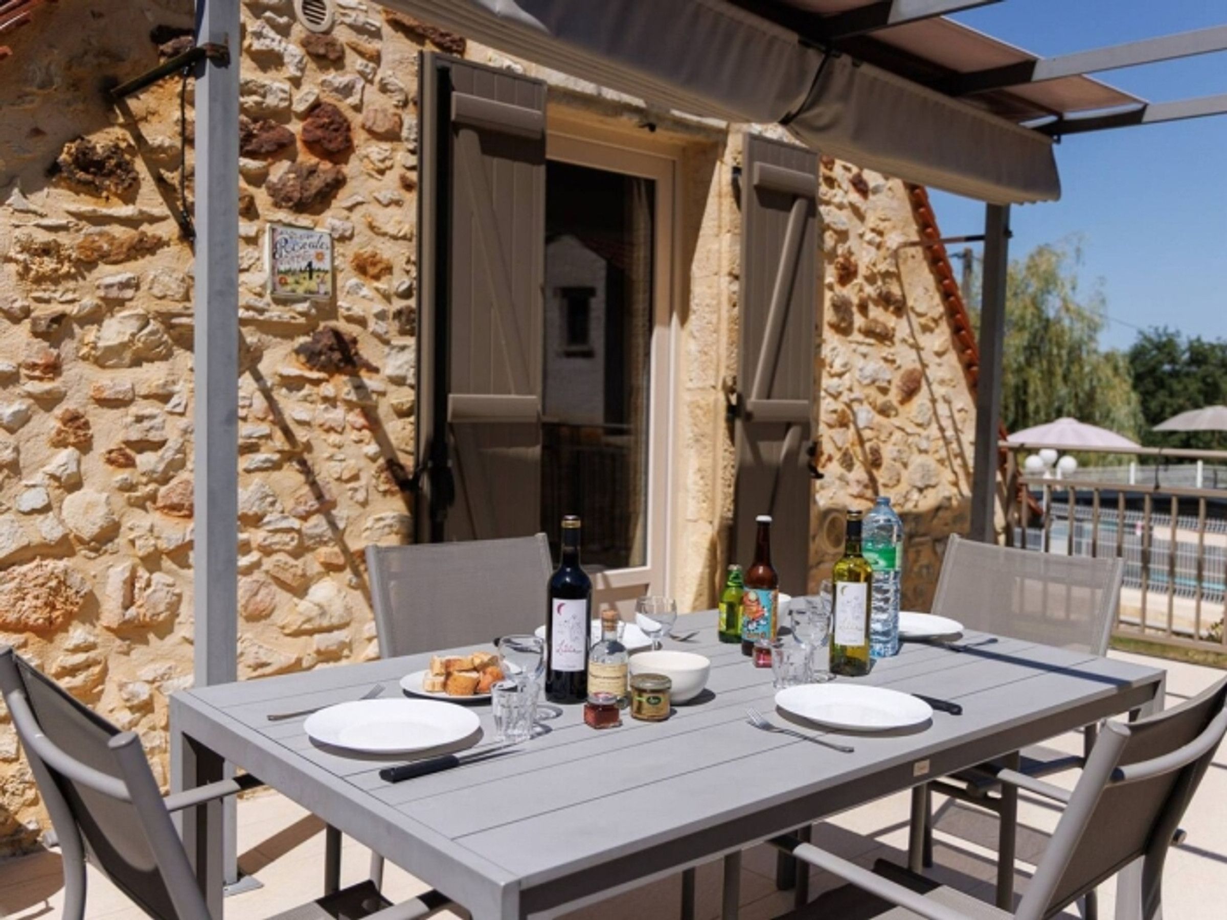 Un espace de restauration en plein air, avec un mur en pierre en arrière-plan, une table dressée avec des assiettes, des couverts et des bouteilles, et une porte entrouverte donnant sur une pièce.