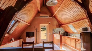 Ein gemütliches Dachzimmer mit schrägen Wänden, einem Esstisch mit bunten Tischdecken, einem Fernseher und einer Küchenzeile mit Holdekor und einem Kühlschrank.