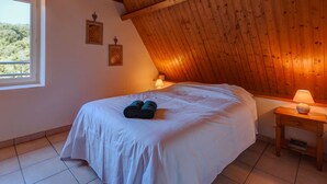 Ein Schlafzimmer mit schrägen Wänden, Holzbalken, einem Bett mit weißer Bettwäsche, zwei an der Wand befestigten Bilderrahmen und einem Nachttisch mit Lampe.