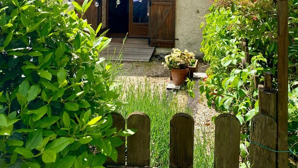 Une maison de campagne avec une cheminée, des portes en bois et un jardin où poussent différentes plantes, le tout entouré d’une clôture en bois.