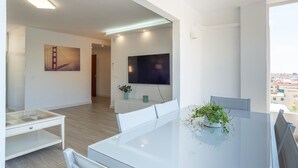 Een moderne woonkamer met een glazen tafel, witte meubels, een flatscreen televisie en een schilderij van een brug.