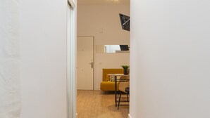 Un couloir mène à une pièce où se trouvent un canapé jaune, une petite table et une horloge au mur.
