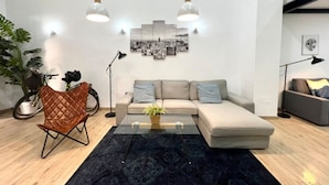 Uma sala de estar moderna com um sofá seccional, uma mesa de centro de vidro e um tapete azul escuro. Há duas luminárias pendentes sobre o sofá e uma obra de arte com paisagem urbana na parede.