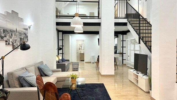 Uma sala de estar ampla com um sofá, poltronas e uma mesa de centro. Uma escadaria leva a um andar superior.