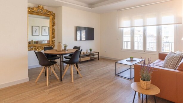 Ein modernes Wohnzimmer mit einem Holzessttisch, schwarzen Stühlen, einer Couch und einem an der Wand befestigten Fernseher.