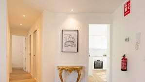 Ein Flur mit Holzboden, einem gerahmten Bild an der Wand, einem goldfarbenen Konsolentisch und einem sichtbaren Küchenbereich mit weißen Schränken und einem roten Feuerlöscher.