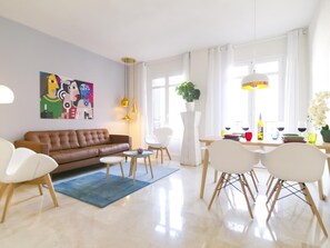 Ein modernes Wohnzimmer mit einem braunen Ledersofa, weißen Stühlen, einem Esstisch mit Stühlen, einem blauen Teppich und einem farbenfrohen abstrakten Gemälde an der Wand.