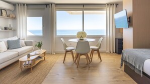 Ein modernes Hotelzimmer mit einer Sitzecke, einem runden Esstisch mit Stühlen, einem an der Wand montierten Fernseher und großen Fenstern mit Meerblick.