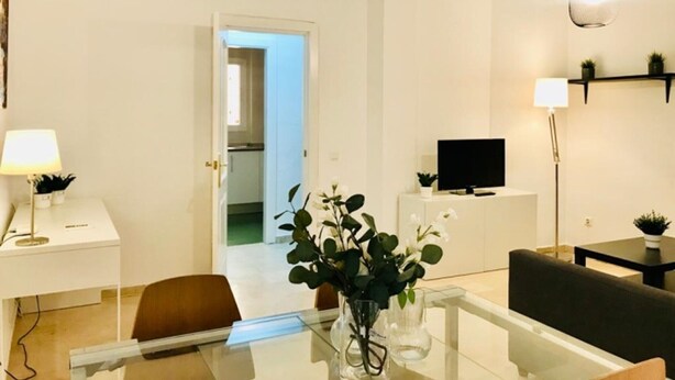 En moderne stue med et glasbord, en sofa, et fjernsyn på et hvidt skab og to hængelamper.