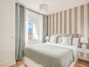 Ein Schlafzimmer mit einem Bett, zwei Nachttischen, einem Fenster mit Vorhängen und Blick auf Gebäude.