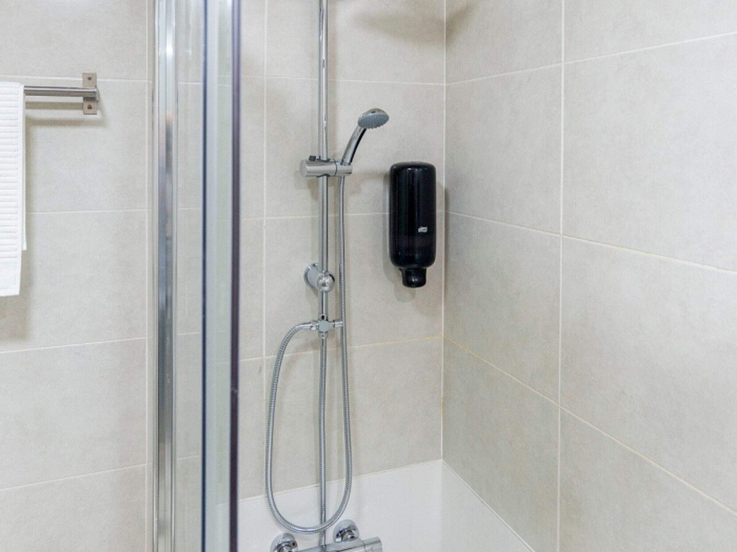 Dusche mit Glastür, Handbrause und Seifenspender an der Wand.