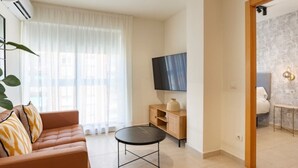 Uma sala de estar moderna com um sofá, uma mesa redonda pequena, uma televisão na parede e uma janela grande com cortinas leves.