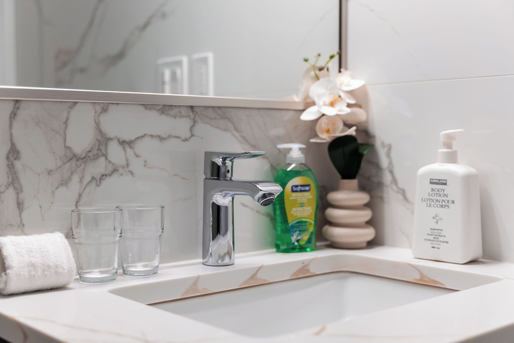 Une salle de bain avec un lavabo en marbre, deux verres transparents, une serviette blanche, un distributeur de savon vert et un flacon de lotion blanche.