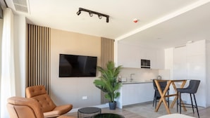Un soggiorno moderno con un letto grande, una TV a parete, una zona relax con una poltrona in pelle marrone e un angolo cottura con tavolo e sedie.