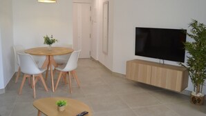 Ein modernes Wohnzimmer mit einem runden Holzesstisch, weißen Stühlen, einem an einer Holzkommode befestigten Flachbildfernseher und einem kleinen Topfpflanzen auf dem Boden.