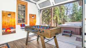 Une pièce avec un baby-foot en bois, une fenêtre donnant sur les arbres et un jacuzzi à l’extérieur.