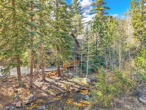 A cabin surrounded by dense forest, a wooden deck, and a small stream.
