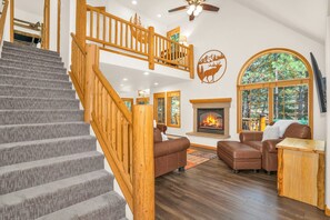 A spacious living area with a staircase, a fireplace, and a large window with a view of trees.