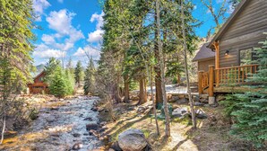 Une cabane dotée d’une terrasse en bois, bordée d’un ruisseau et entourée d’arbres.