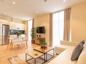 Un salon moderne avec un canapé, une table basse et un téléviseur. On y trouve également un coin repas avec des chaises et une table, ainsi qu’une cuisine équipée.