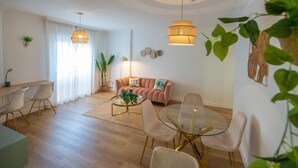 Ein modernes Wohnzimmer mit einer Couch, Sesseln, einem Couchtisch und einem Essbereich mit einem Glastisch und Stühlen. Es gibt Pflanzen, eine Lampe und eine Deckenleuchte.
