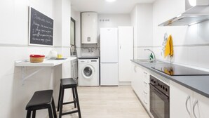 Een moderne keuken met witte kasten, een ingebouwde wasmachine, een spoelbak en een fornuis. Er staan twee zwarte krukken en een prikbord met tekst aan de muur.