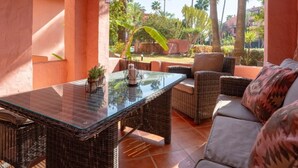 An outdoor patio with wicker furniture, a glass-top table, and a view of palm trees and a garden. - Elegant Marbella Apartment with Direct Pool Access and Parking (Marbella)