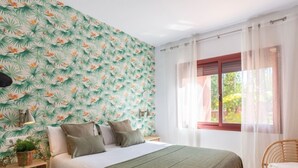 A bedroom with a bed, a nightstand, a chair, a window with curtains, and a wall with a tropical leaf pattern. - Elegant Marbella Apartment with Direct Pool Access and Parking (Marbella)