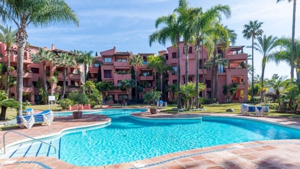 A hotel with a swimming pool surrounded by palm trees and a red building facade. - Elegant Marbella Apartment with Direct Pool Access and Parking (Marbella)
