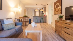 A modern living room with a sofa, a coffee table, and a TV on a wooden cabinet. There is a dining area with a table and chairs, and a kitchen in the background. - Elegant Marbella Apartment with Direct Pool Access and Parking (Marbella)