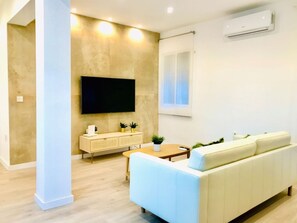 Un salon moderne avec un canapé beige, un téléviseur à écran plat fixé au mur, une petite table sur laquelle est posée une plante en pot et une fenêtre avec des rideaux.
