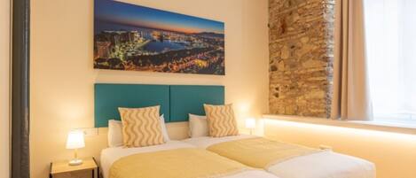 Un lit double avec une tête de lit de couleur turquoise, une table de chevet avec une lampe et un tableau représentant un paysage urbain accroché au mur.