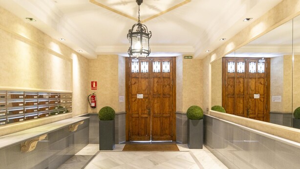 Un vestíbulo de hotel con piso de mármol, puertas de madera, un extintor, un letrero de salida de incendios y un espejo grande que refleja las puertas.