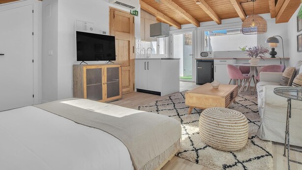 Ein modernes Interieur mit Holzboden, einem Fernseher auf einer Kommode, einer Küchenzeile und einer Sitzecke mit Tisch und Stühlen.