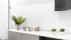 Un salon moderne avec un meuble TV blanc, une plante en pot et des lettres décoratives formant le mot « Bienvenue ».