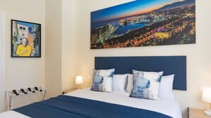 A hotel room with a large bed, a nightstand, a lamp, and a painting of a cityscape. - Charming apartment near La Malagueta, Malaga (Málaga)