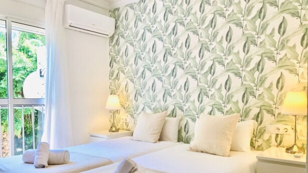 A hotel room with two beds, a window with curtains, an air conditioning unit, and a wall with a leafy pattern. - Spacious apartment with balcony in Málaga (Málaga)