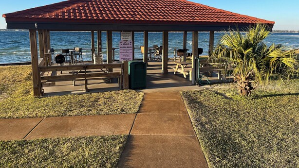 Ein Gazebo mit Ziegeldach und Blick auf ein Gewässer, ausgestattet mit Picknicktischen und -stühlen, einem Mülleimer und einem Schild mit Regeln.