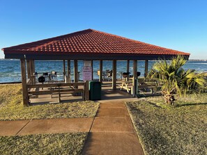 A gazebo with a tiled roof overlooking a body of water, featuring picnic tables and chairs, a trash can, and a sign with rules. - 1-bedroom condo on Navarre Beach with private beach access, pier, and pavilion. (Navarre)
