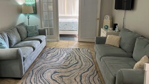 Ein Wohnzimmer mit zwei grauen Sofas, einem gemusterten Teppich, einem Fernseher und einem Schild mit Strandmotiv.