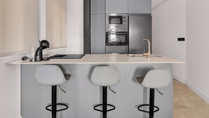 Una cocina moderna con horno empotrado, microondas y refrigerador. Hay tres taburetes de bar y un lavamanos con grifo.